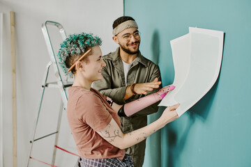 Young adult woman with short curly hair and tattoos holding paint samples, smiling and discussing renovation plans with young adult man in casual clothing near teal wall