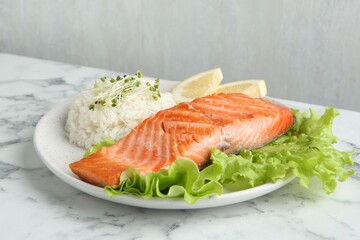 Piece of tasty grilled salmon, lemon, lettuce, rice and microgreens on light marble table against grey background, closeup