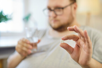 Obese man holding pill and water glass to take medicine for routine treatment at home. Hand of...