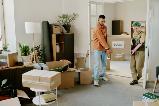 Young adult man and young adult woman carrying cardboard box labeled books while moving into new home, surrounded by unpacked boxes and houseplants