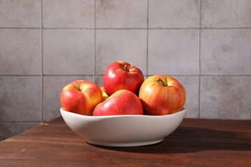 Ripe red apples in bowl on wooden table near grey tiled wall, closeup