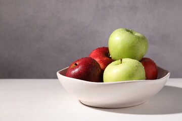 Fresh red and green apples with water drops in bowl on white table against grey background, closeup. Space for text