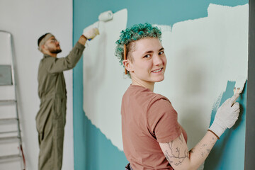 Portrait of young adult Caucasian woman smiling at camera while painting wall with roller, young adult man with beard and glasses painting wall in background during home renovation