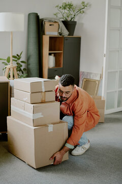 Young adult man lifting cardboard boxes in living room during moving and renovation process, focusing on organizing belongings and preparing new home environment