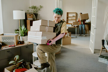 Young adult Caucasian woman with prosthetic arm carrying stack of cardboard boxes while moving into...