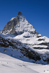 Iconic Matterhorn peak covered in winter snow, rising sharply against a clear deep blue sky. Dramatic alpine landscape with rugged cliffs and glacier textures.