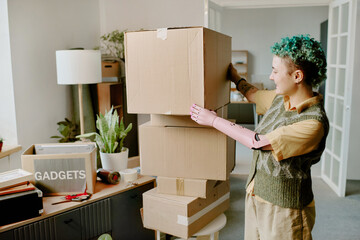 Young adult Caucasian woman with prosthetic arm stacking cardboard boxes in living room during moving and renovation, visible plants and packed items indicating relocation process