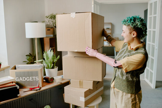 Young adult Caucasian woman with prosthetic arm stacking cardboard boxes in living room during moving and renovation, visible plants and packed items indicating relocation process