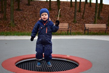 Toddler boy in a blue winter overall jumping on an in-ground playground trampoline. Behind him are park benches, autumn trees and soft rubber flooring on a cloudy day.