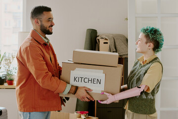 Young adult man handing moving boxes labeled kitchen to young adult woman with prosthetic arm, both standing in home interior during renovation and relocation