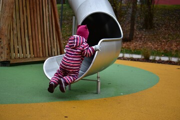 Toddler girl in a striped winter overall climbing up a metal playground slide from the bottom. Bright rubber flooring and wooden structures create a colorful, modern outdoor play scene.