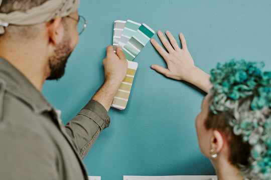Young adult man and young adult Caucasian woman comparing paint color swatches against wall, preparing for home renovation, both focusing on selecting suitable shade together - Powered by Adobe