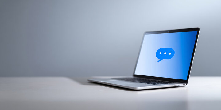 A laptop with a speech bubble on the screen is sitting on a white table against a grey background - Powered by Adobe