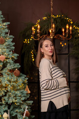 Fototapeta premium Smiling pregnant woman in a striped sweater gently holding her baby bump, posing by a decorated christmas tree and a festive arch with a chandelier