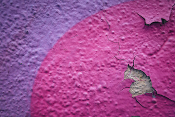 Close-up view of textured building wall with peeling paint. The surface features vibrant pink and purple colors, revealing a rough, damaged area underneath.