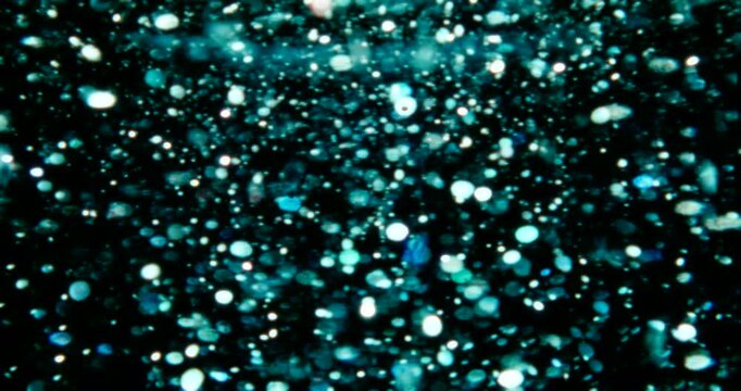 Particles float in water over a dark background, reflecting top light in green and blue hues, creating a glittering, halo-like, cosmic universe effect.