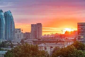 Vibrant Kyiv cityscape at sunset, with multiple high-rise buildings silhouetted against an orange and pink sky Some structures are illuminated, contrasting the urban landscape with a lush park in th