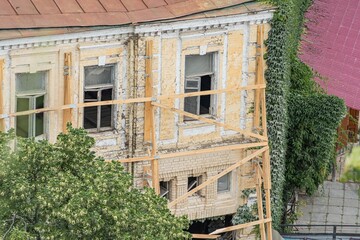 Old European city building renovation in progress Unfinished facade with scaffolding, wooden panels, muted yellow and gray color palette Some intact windows, others boarded up Traditional window d