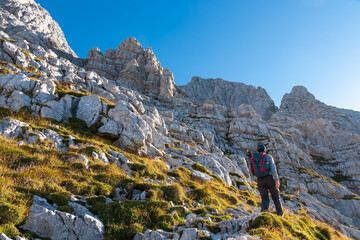 Naklejka premium A hiker equipped with a helmet and backpack ascends a steep rocky slope, gazing at the towering limestone cliffs of Jof di Montasio in the Julian Alps, Friuli Venezia Giulia, Italy, on a sunny day.