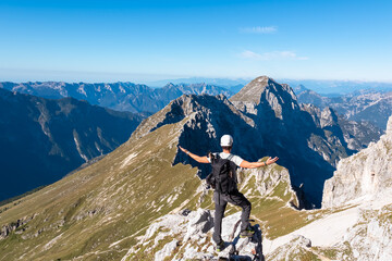Fototapeta premium A climber with a backpack stands with arms wide open on the jagged summit of Jof di Montasio. He embraces the panoramic view of the Julian Alps in Friuli Venezia Giulia, Italy during a sunny hike.
