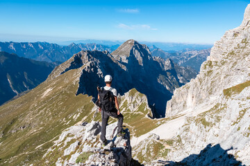 A male hiker stands on a rocky ledge admiring the sheer cliffs of the Julian Alps near Jof di Montasio. The rugged mountain landscape of Friuli Venezia Giulia, Italy stretches to the horizon.