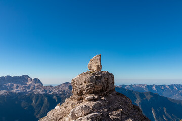 A solitary rock cairn stands prominently atop the rugged summit, overlooking the breathtaking panorama of the Julian Alps near Jof di Montasio in Friuli Venezia Giulia, Italy, under a clear blue sky.