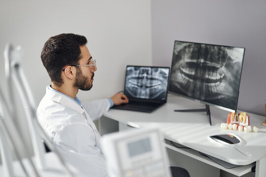 Middle Eastern man wearing lab coat analyzing dental X ray images on computer monitor and laptop in dental office, dental model samples placed on desk, attending patient care