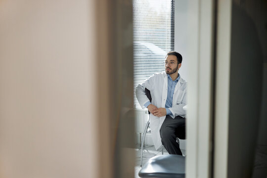 Young dentist wearing lab coat sitting in dental office looking to side, appearing thoughtful, modern clinic environment visible through partially open door