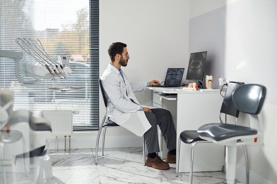 Middle Eastern man wearing lab coat sitting at desk in dental office, reviewing dental x rays on computer monitor with dental model and dental equipment visible nearby