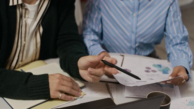 Close up of unrecognizable coworkers sitting at round table, reading report including analytics, schemes and charts