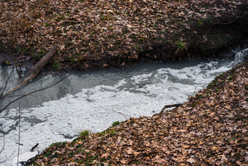 Polluted stream with dirty gray water and foam on the surface, surrounded by brown fallen leaves and muddy banks in an autumn forest. Environmental pollution and ecological damage concept