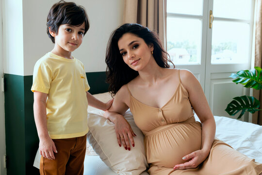 Pregnant young woman sitting on bed holding belly, looking at camera, with boy child standing beside her, both posing in bright bedroom interior - Powered by Adobe