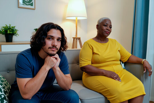 Portrait of young man sitting on sofa leaning forward with hands under chin next to adult Black woman sitting on sofa looking at camera