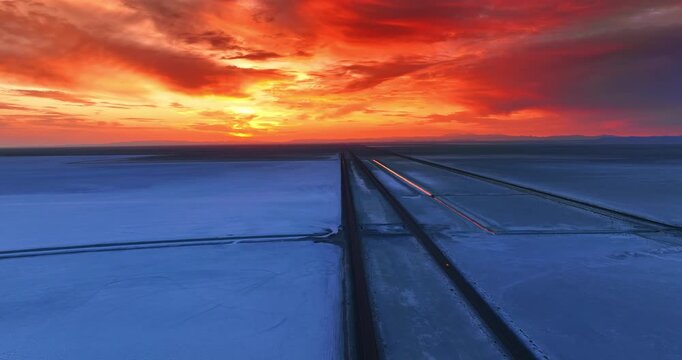 Not many cars go by two parallel straight roads through the Bonneville Salt Flat, Utah, United States. The cloudy sky is orange from the setting sun.