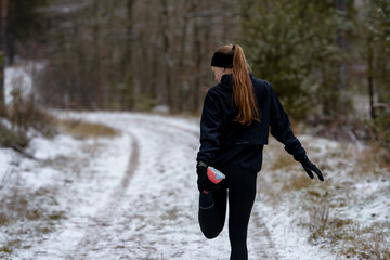 Female figure in athletic leggings and jacket maintaining her routine. She stops to stretch her muscles on a snow-covered forest path during training.