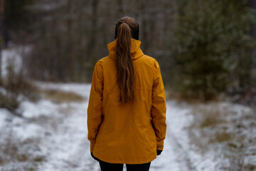 Young woman in a bright yellow jacket stands alone on a cold, snow-dusted forest path, looking forward. This image captures solitude, winter exploration, and outdoor adventure. © Alina Mosinyan