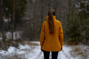 View from behind of a woman with long hair in a high ponytail, wearing an oyellow rain jacket in a wintry forest landscape. Ideal for themes of travel, hiking, and seasonal fashion.