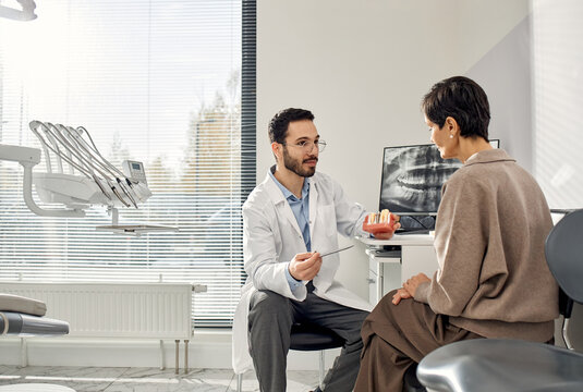 Middle Eastern male dentist explaining dental procedure, using jaw model to Caucasian patient during consultation in dental office with dental equipment visible
