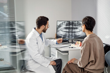 Young male dentist explaining dental x ray results to middle aged Caucasian woman patient in dental...