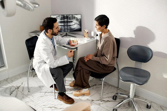 Middle Eastern dentist explaining dental procedure to middle aged Caucasian patient in dental office, both sitting and discussing treatment plan with dental x-ray visible in background - Powered by Adobe