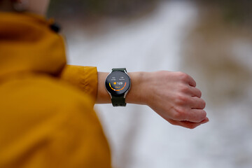 Close-up of an athlete checking heart rate on a smartwatch during a winter workout. The digital screen displays 129 bpm and Zone 2 intensity. Person wears a yellow jacket, snowy outdoor environment