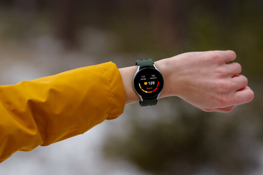 Close-up of an athlete checking heart rate on a smartwatch during a winter workout. The digital screen displays 129bpm and Zone 2 intensity. Person wears a yellow jacket, snowy outdoor environment