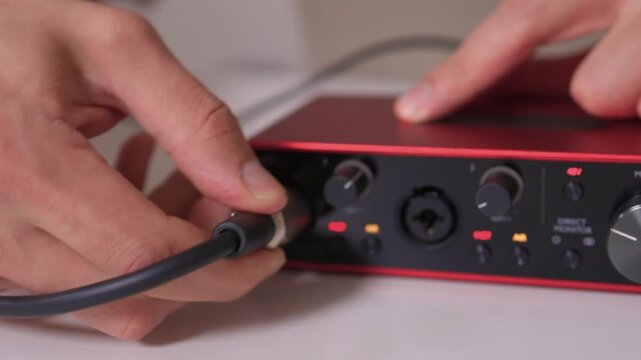 Hands plug XLR cable into interface, closeup red external sound card on white desk, careful insertion of metal connector, studio recording setup for podcasting and streaming
