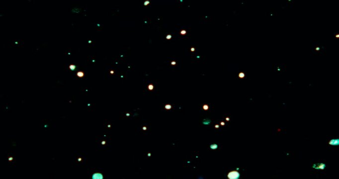 Particles float in water over a dark background, reflecting top light in green and blue hues, creating a glittering, halo-like, cosmic universe effect.