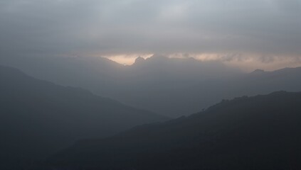 A view of mountain ranges covered in fog and clouds with a faint glow of light breaking through above