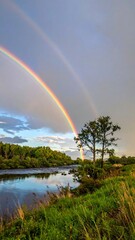 Naklejka premium Vibrant Double Rainbow Over Tranquil River Landscape with Lush Greenery.