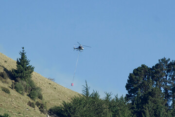 A helicopter carrying water to fight a blaze in pine trees near Goodwood, Otago NZ. Goodwood is a small coastal settlement on New Zealand's South Island.