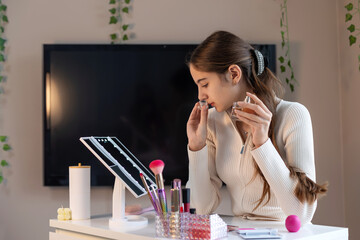 Teen girl testing fragrance at makeup table. A young girl in a cream top gently smells a perfume bottle while seated at a vanity table with beauty products and a lit mirror.