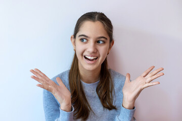 Excited teenage girl with surprised expression. Teen girl in gray sweater smiling with wide eyes and raised hands, expressing surprise or excitement against a plain background.