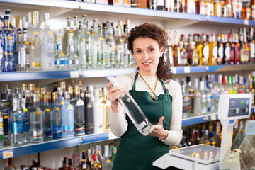 Positive female sales consultant in apron stands near alcohol display case in Russian store, hold bottle of vodka, read label, studying new arrival of item product